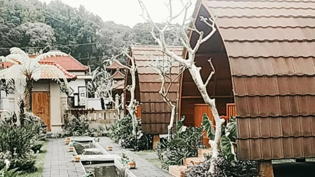 Wooden lumbung cottages with curved shingle roofs beside a garden path and water channel at Umah Lumbung Bedugul