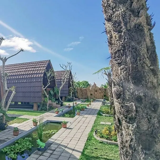 A row of lumbung cottages with brown shingle roofs along a paved garden path under a blue sky